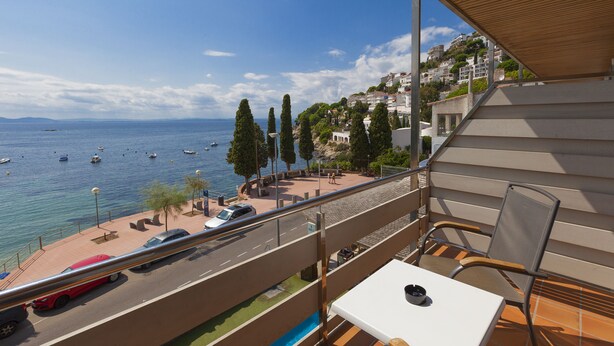 A balcony with a table and chairs, overlooking a coastal road with parked cars and boats on the water. - Canyelles Platja (Roses)