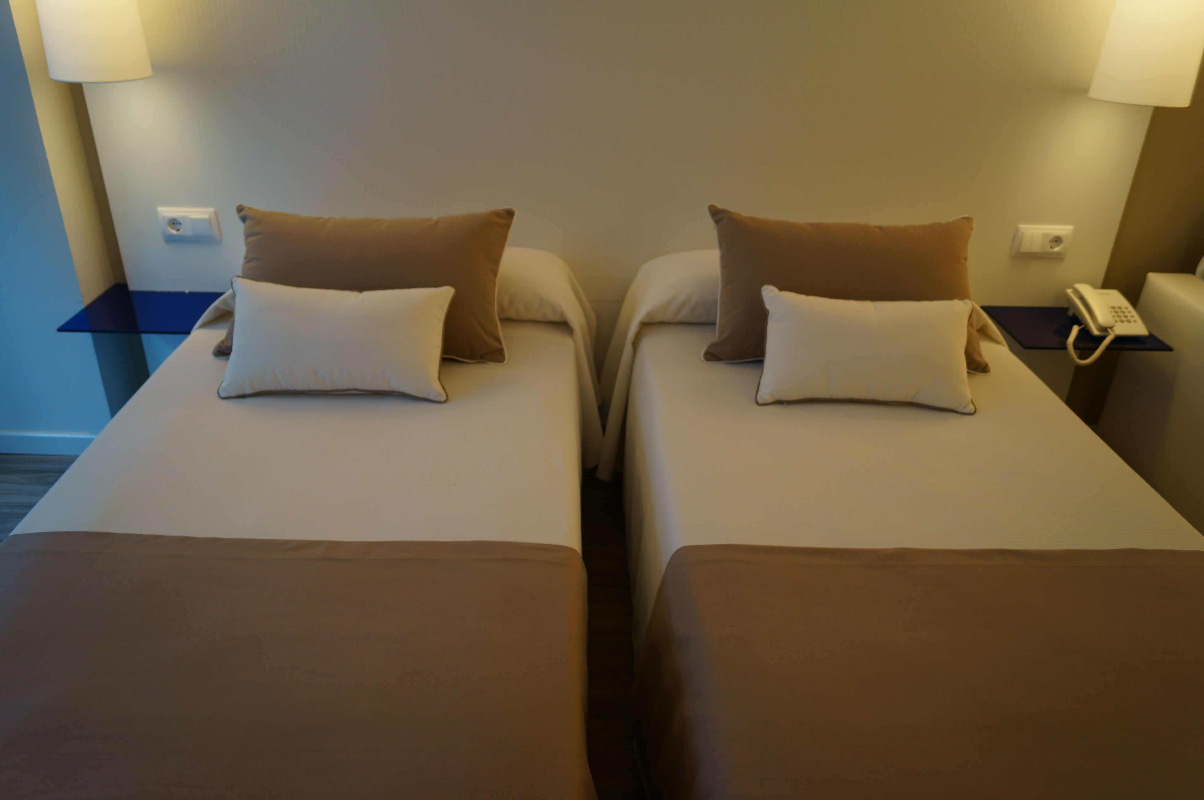 Habitación de hotel con dos camas individuales, cada una con almohada y manta. Se observa un mesita de noche con una lámpara y un teléfono.