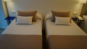 Habitación de hotel con dos camas individuales, cada una con almohada y manta. Se observa un mesita de noche con una lámpara y un teléfono.