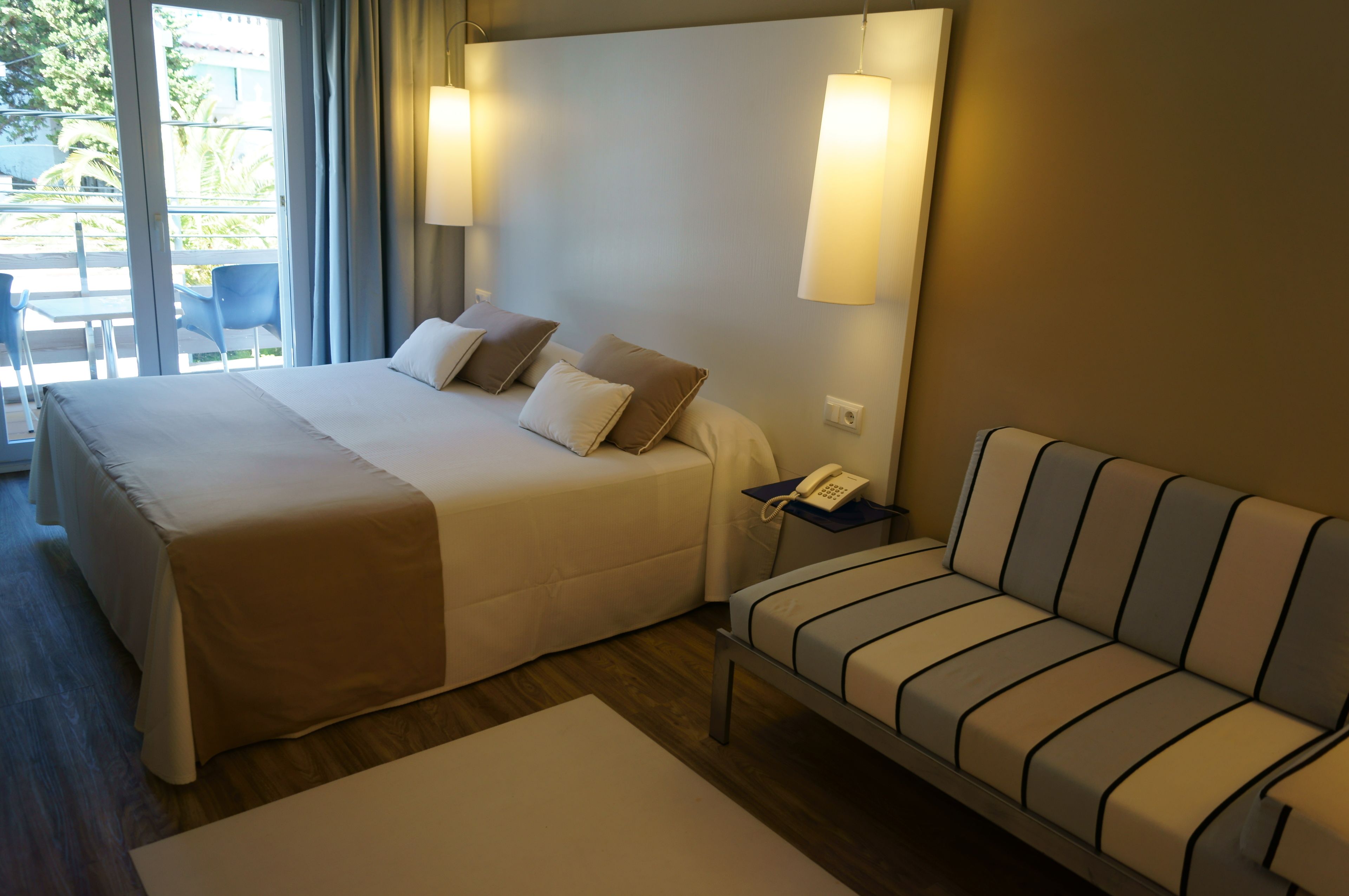 A hotel room with a large bed, a sofa, a bedside table with a phone, and a view of the outdoors through a sliding door.