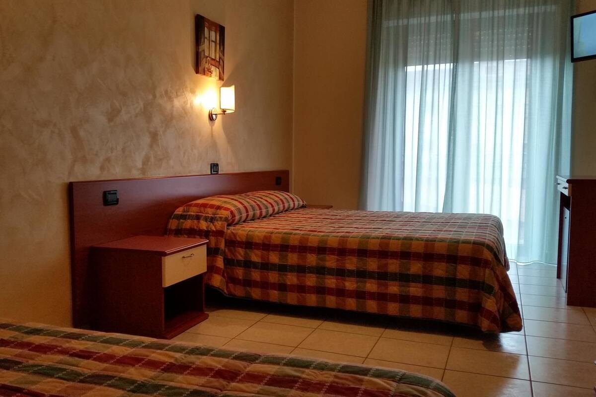 Une chambre d’hôtel avec deux lits, une table de chevet, une lampe fixée au mur et une fenêtre avec des rideaux légers.