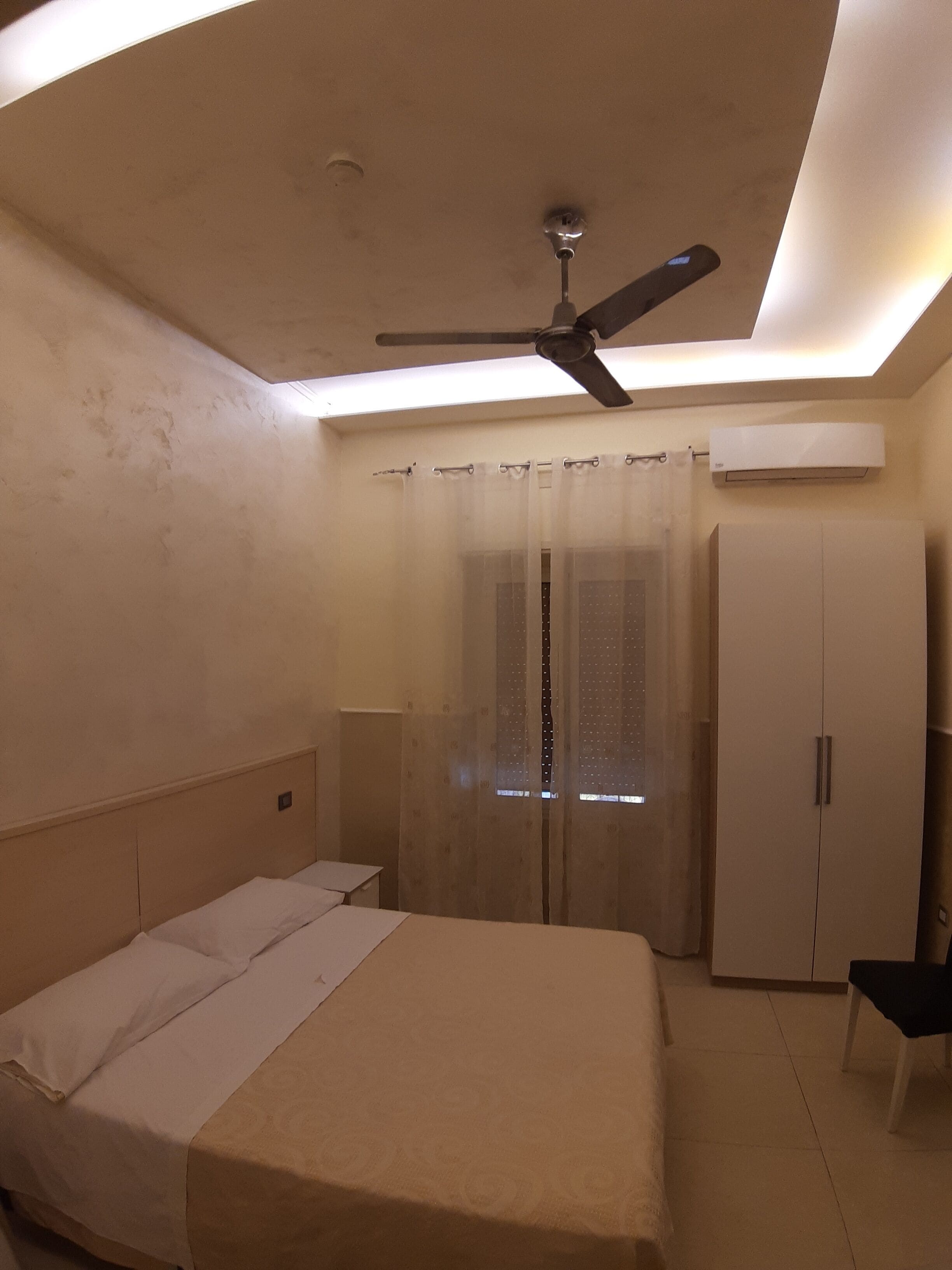 A bedroom with a bed, a ceiling fan, an air conditioning unit, a wardrobe, and a window with curtains.