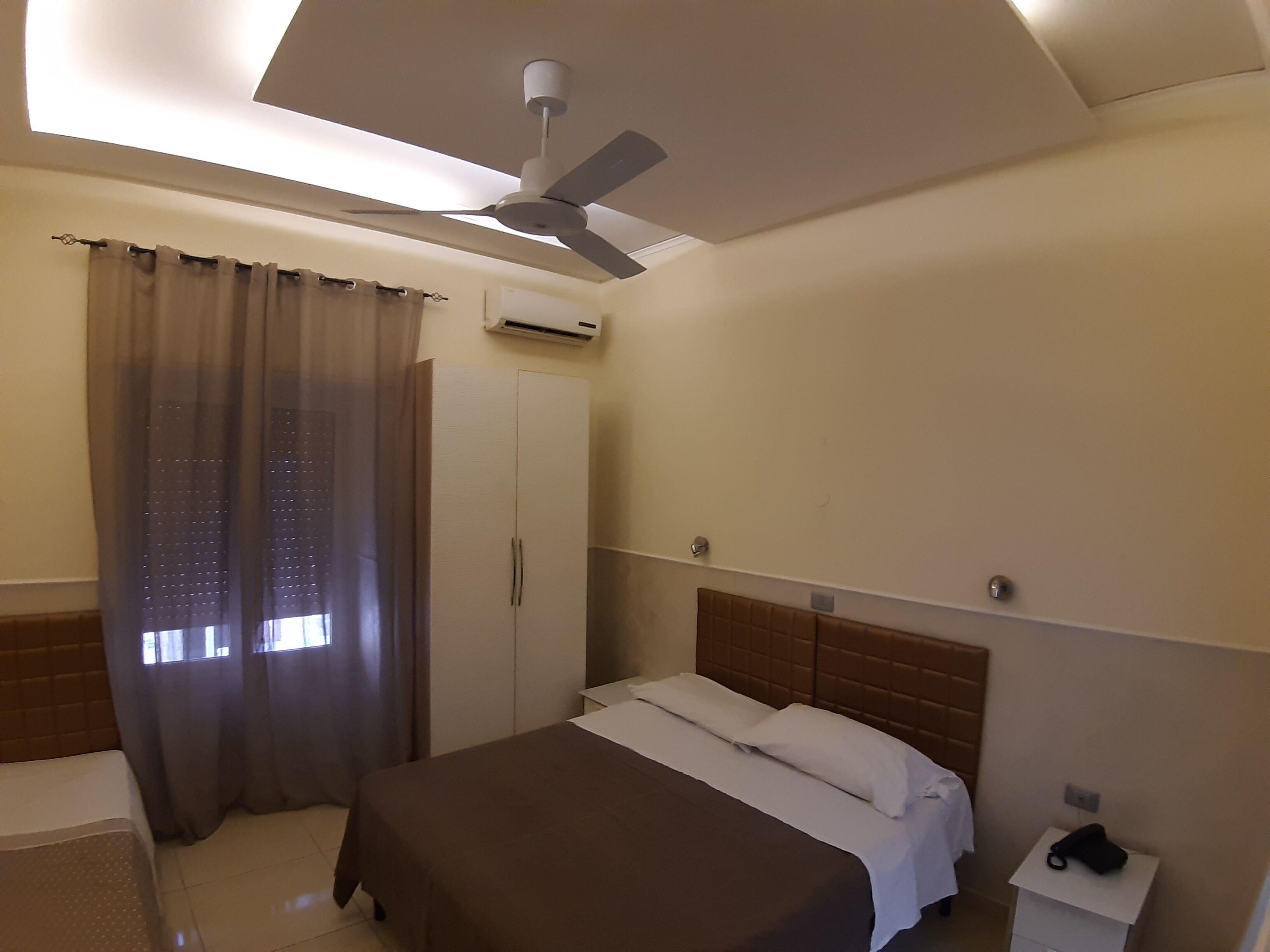 Habitación de hotel con cama, armario, mesita de noche, ventilador de techo y ventana con cortinas.