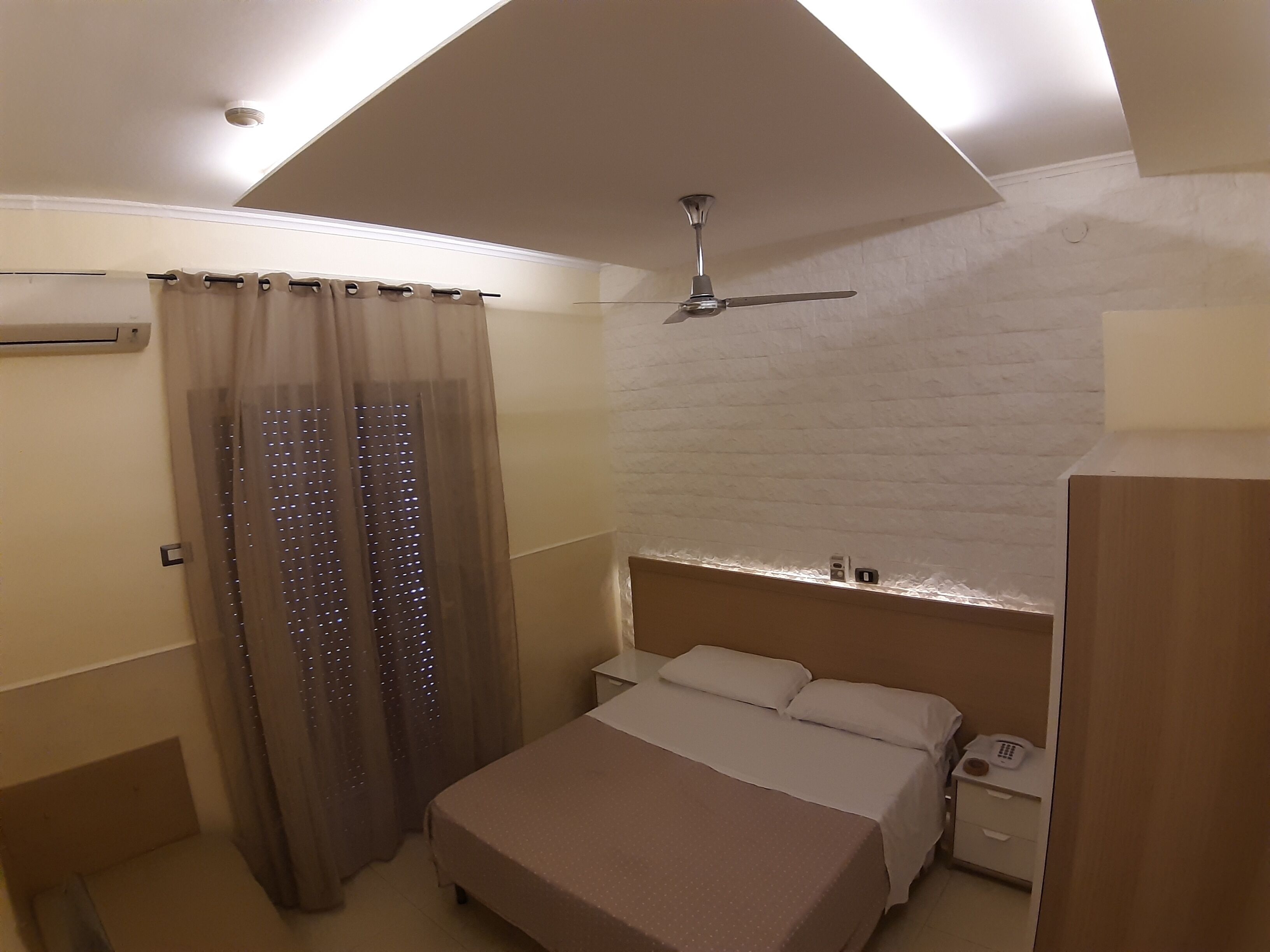 Een slaapkamer met een bed, een plafondventilator, een nachtkastje en een aan de muur bevestigde telefoon.