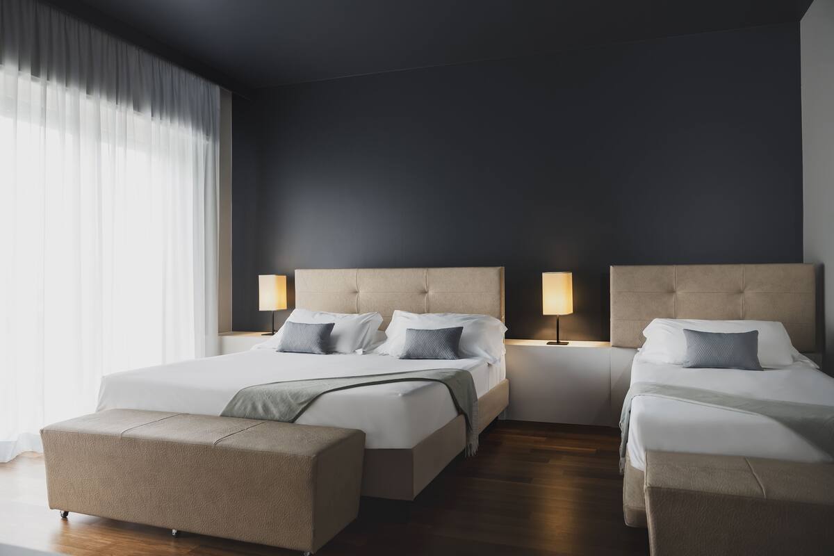 A modern hotel room with two beds, a sofa, and a large window with curtains.
