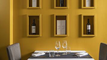 A dining table set with wine glasses, surrounded by chairs, against a yellow wall with wine bottles displayed in shelves.