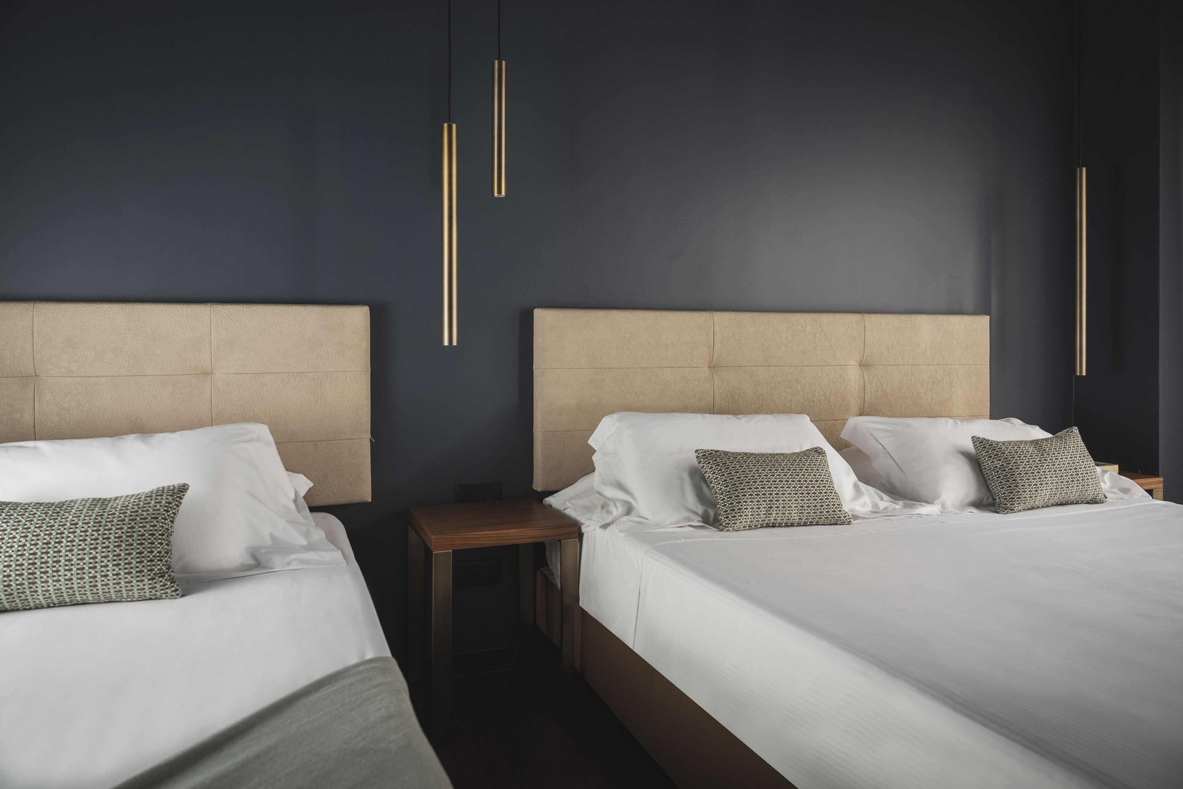 Una camera d'albergo con due letti, ognuno con testiera, biancheria da letto bianca e cuscini decorati. Sono presenti comodini con lampade.