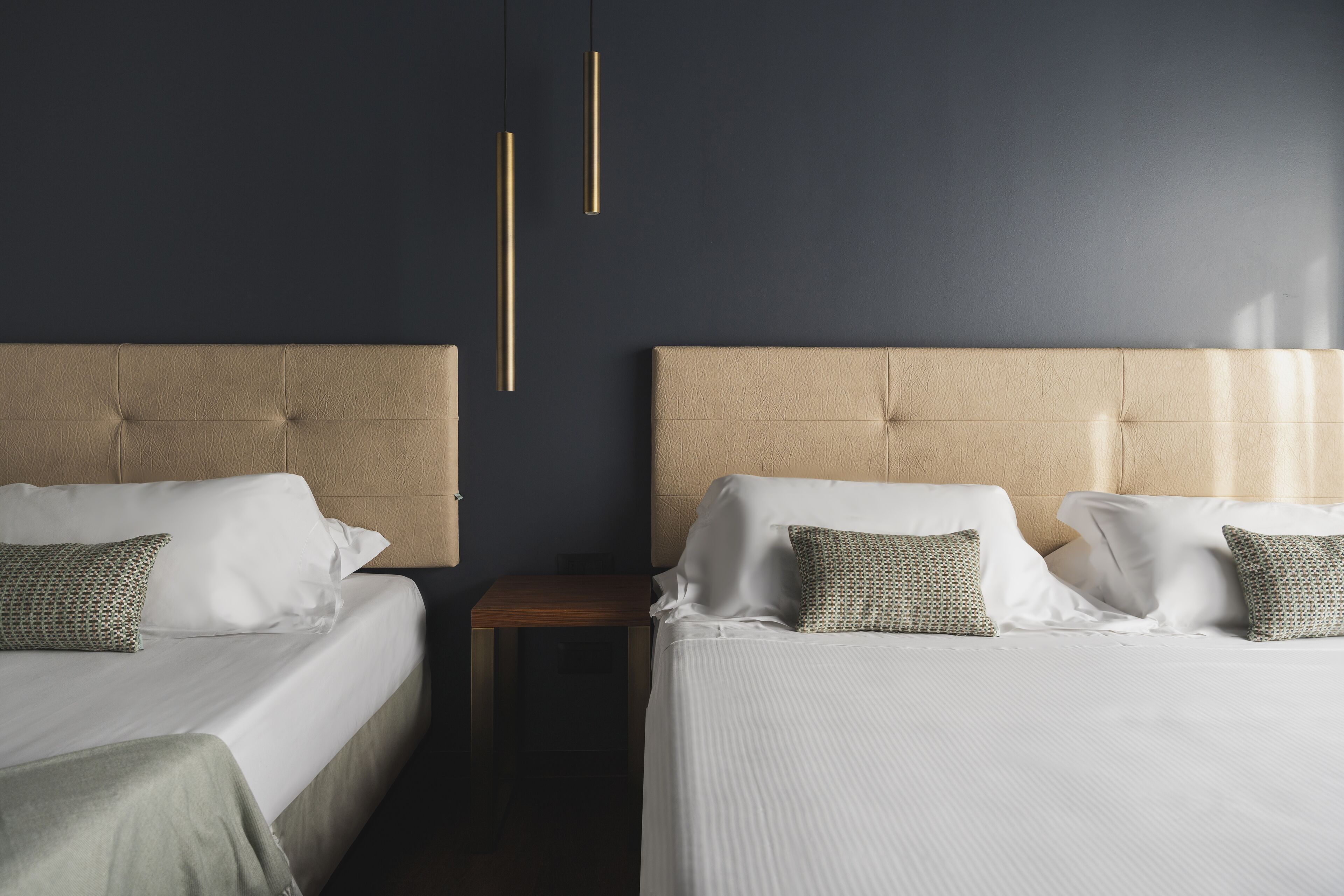 A hotel room with two beds, each with a beige headboard, white linens, and patterned pillows. There is a small wooden nightstand between the beds.