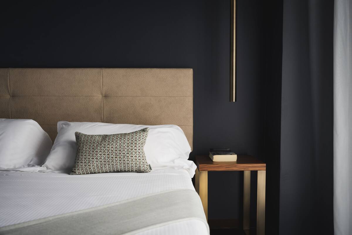 A neatly made bed with white linens and a patterned pillow, a wooden bedside table with a book, and a dark wall with a vertical light fixture.