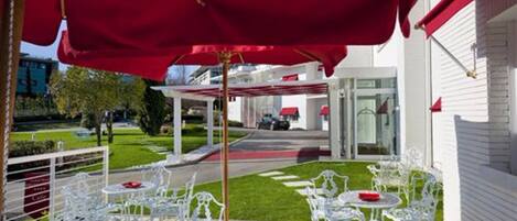 Un pati exterior amb cadires de metall blanc i ombrelles vermelles, un edifici blanc al fons i un jardí ben cuidat.