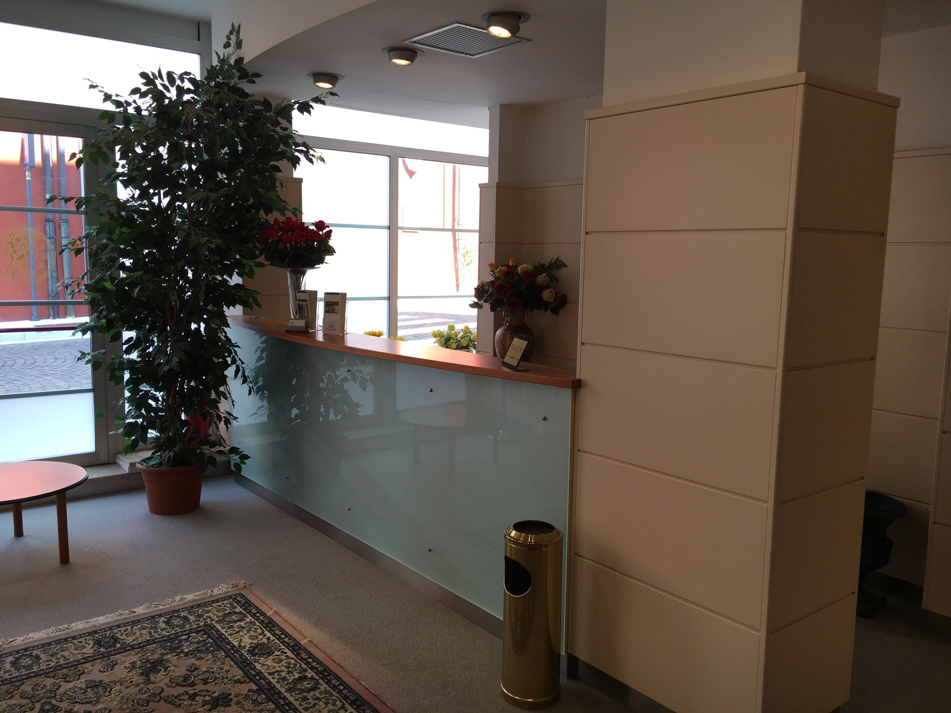 A reception area with a reception desk, potted plants, a trash can, and a view of the outside through large windows.