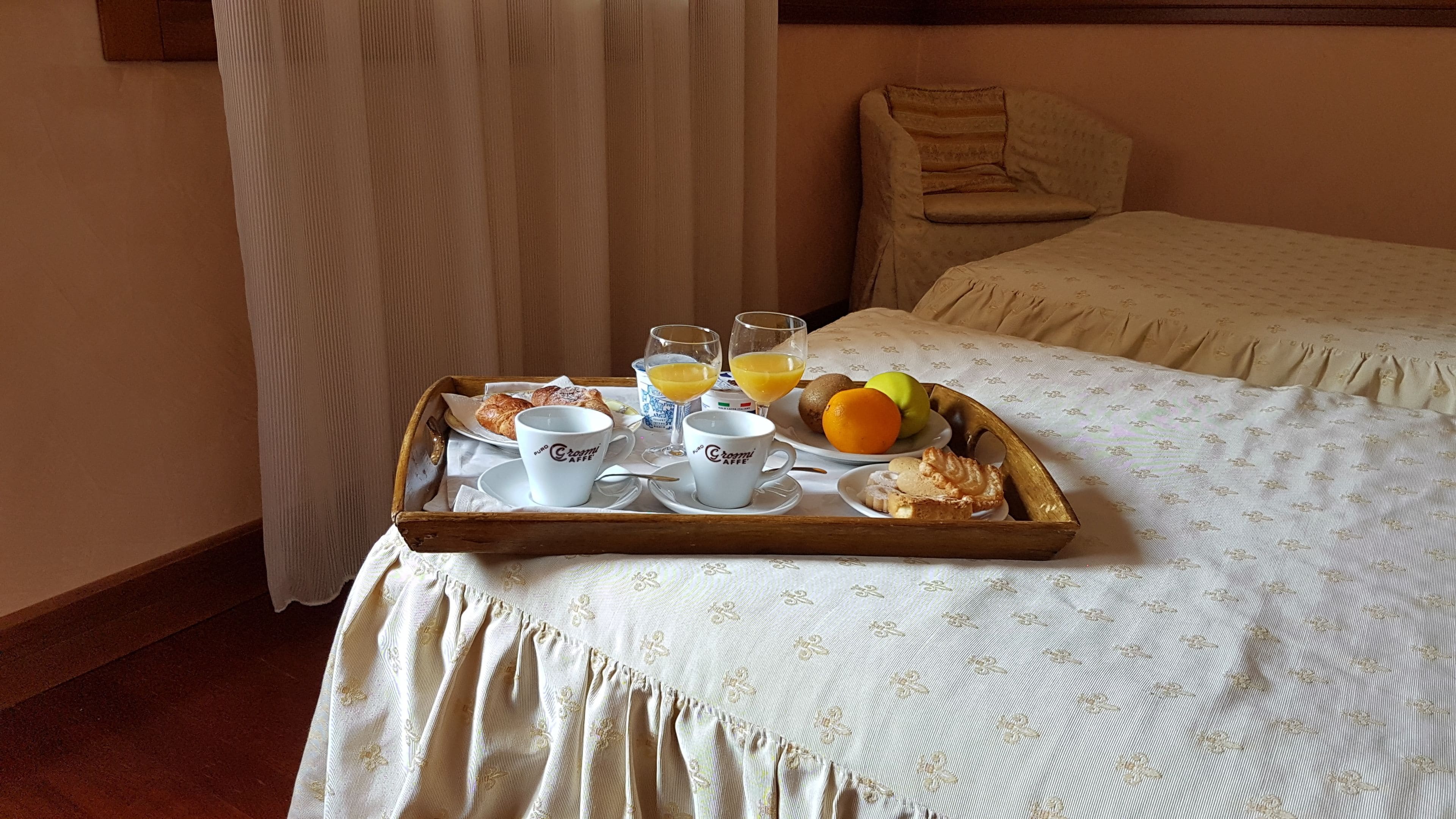 A breakfast tray with croissants, coffee cups, orange juice, and fruit on a bed in a hotel room.