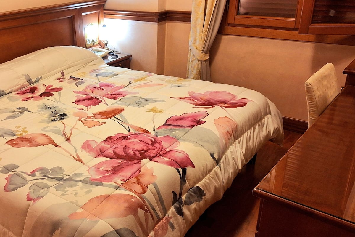 A neatly made bed with a floral bedspread, a wooden headboard, a bedside table with a lamp, and a chair to the right.