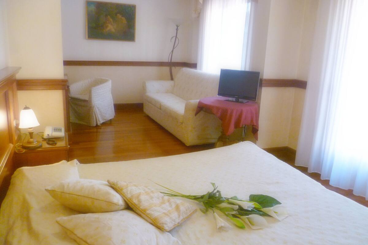 A hotel room with a bed, a sofa, a television on a stand, and a painting on the wall.