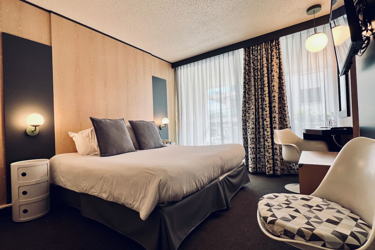 A modern hotel room with a large bed, a desk, and a chair. There are two bedside tables and a lamp on each.