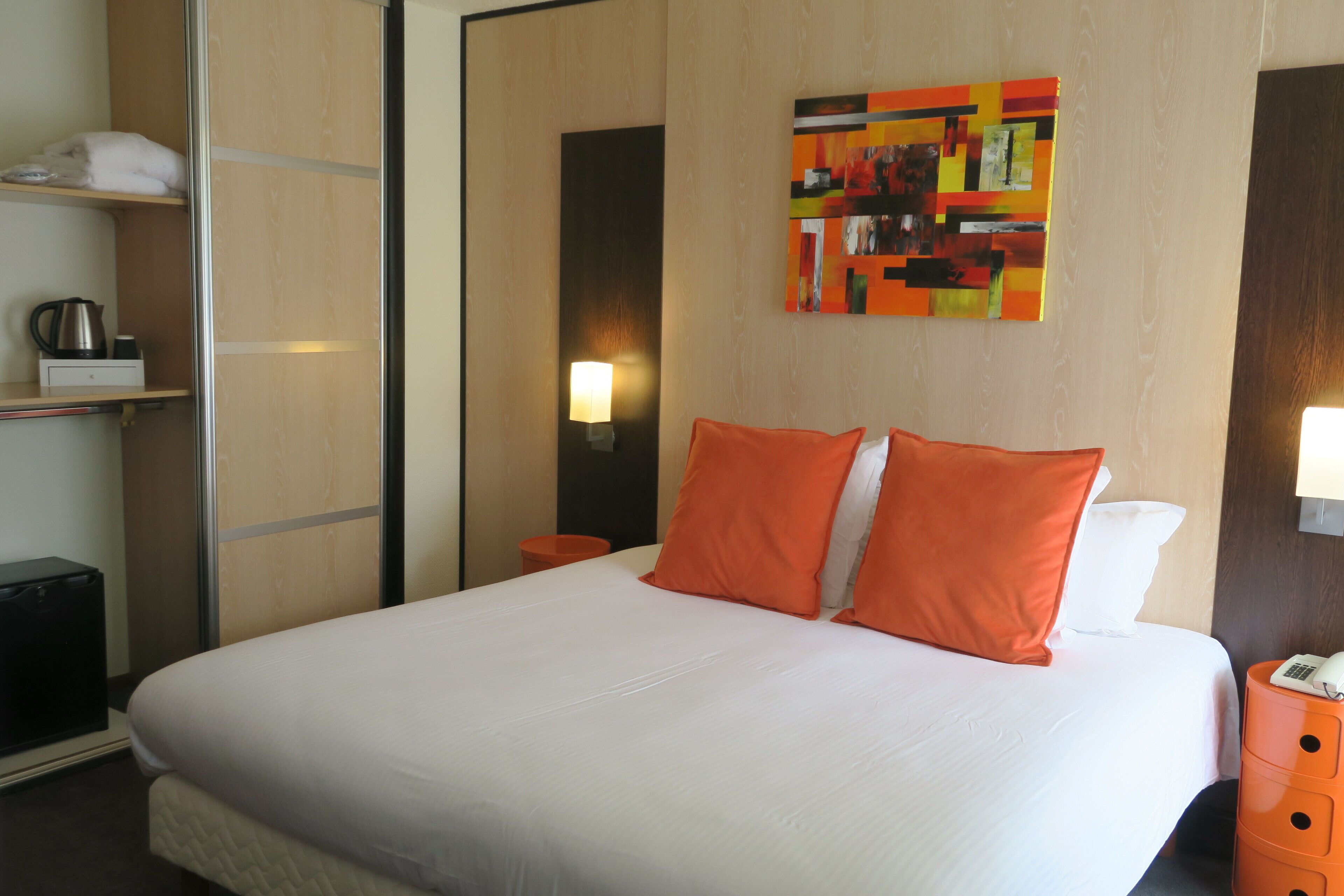 A hotel room with a large bed, orange pillows, a bedside table with a lamp, a telephone, and a wall art piece.