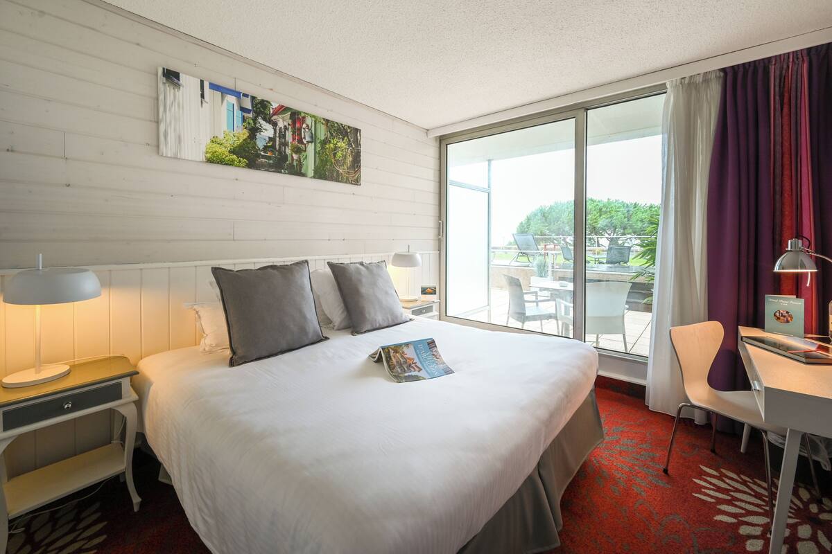 A hotel room with a bed, a desk, a chair, a window with curtains, and a painting on the wall.