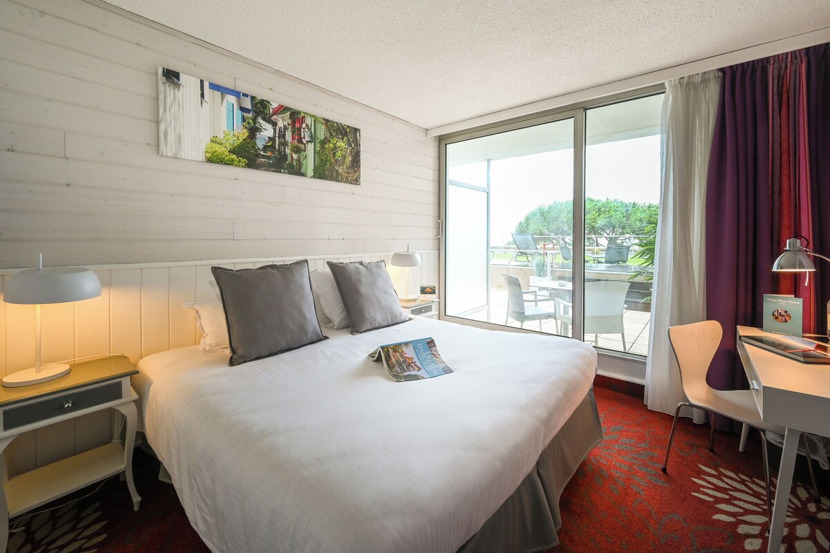 Habitación de hotel con cama, escritorio, silla, ventana con cortinas y un cuadro en la pared.