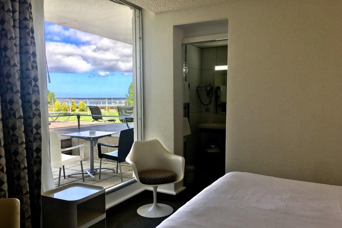 A hotel room with a bed, a small desk, a chair, and a view of the outdoors.