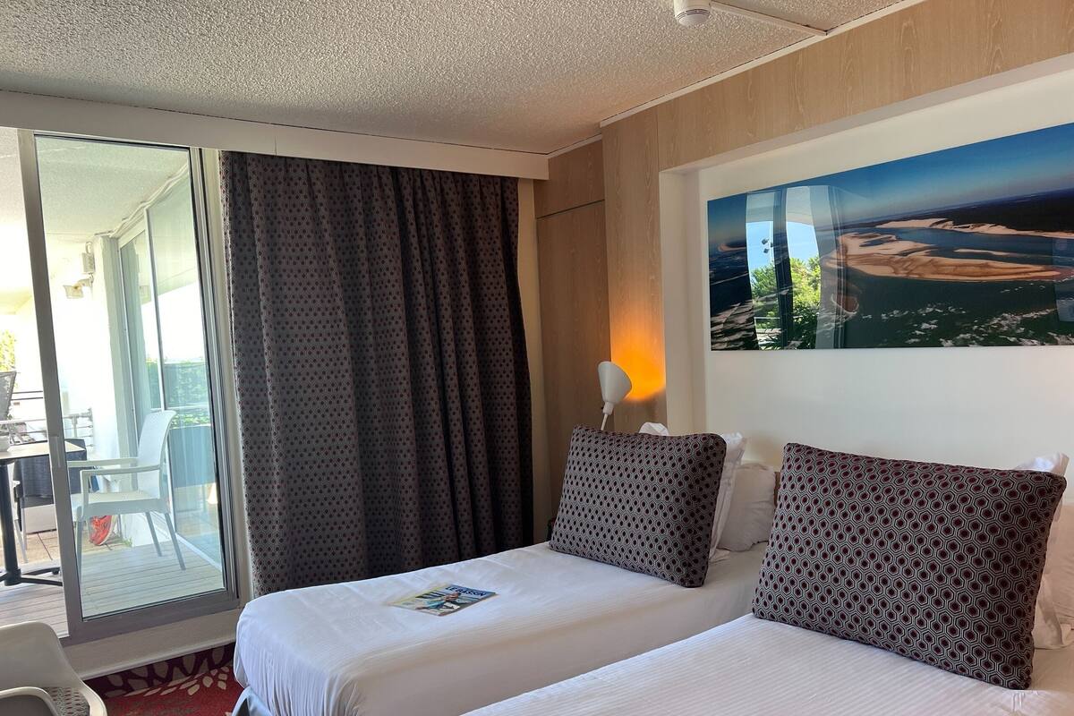 A hotel room with two beds, a large window with curtains, a painting on the wall, and a view of the outdoors.