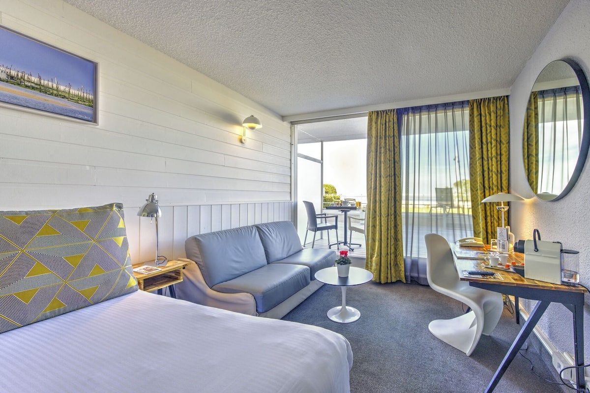A hotel room with a bed, a sofa, a small round table, a desk, a chair, a lamp, and a window with curtains.