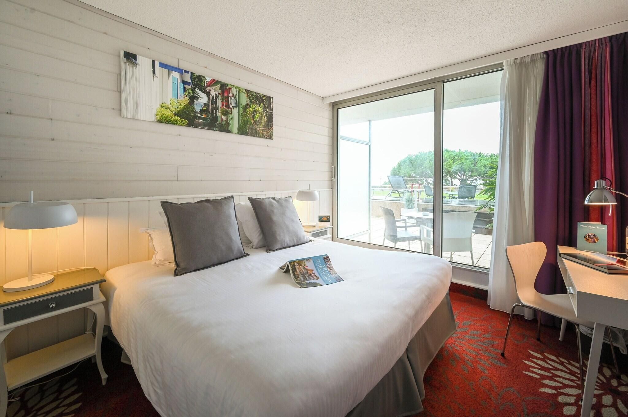 A hotel room with a bed, a desk, a chair, a window with curtains, and a painting on the wall.