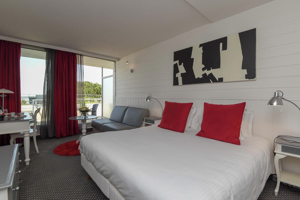 A hotel room with a large bed, a desk, a chair, a small table, and a view of the outdoors.