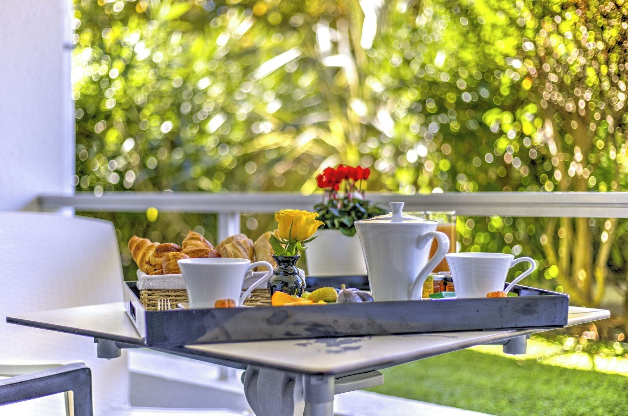 A breakfast tray with croissants, a vase with a yellow flower, and white cups on a balcony with a green garden view.