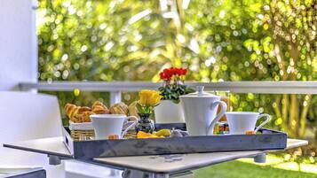 Un plateau de petit-déjeuner avec des croissants, un vase contenant une fleur jaune et des tasses blanches sur un balcon donnant sur un jardin verdoyant.