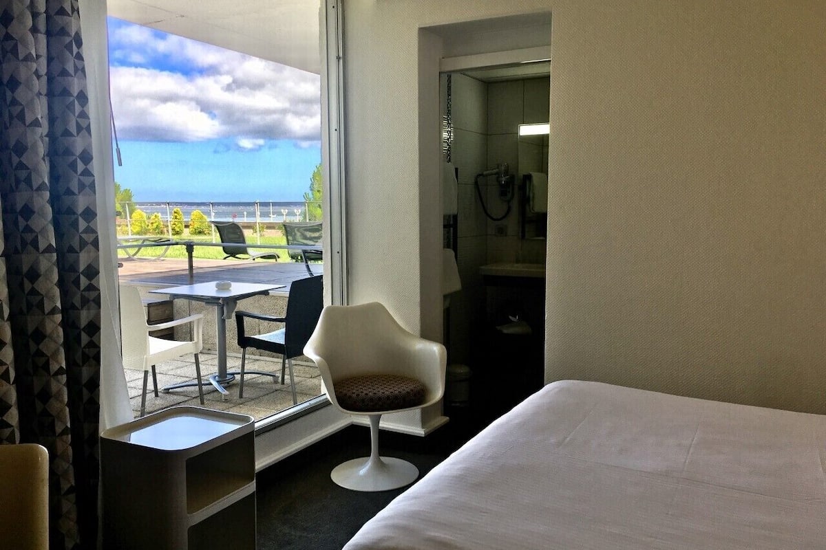 A hotel room with a bed, a chair, a small table, and a view of the outdoors.