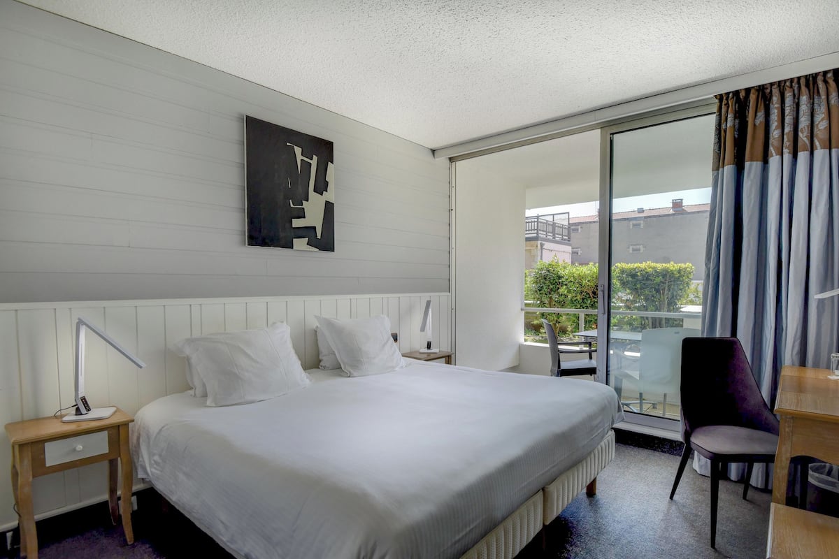 A hotel room with a large bed, a desk, a chair, a nightstand, a lamp, and a sliding glass door to the balcony.