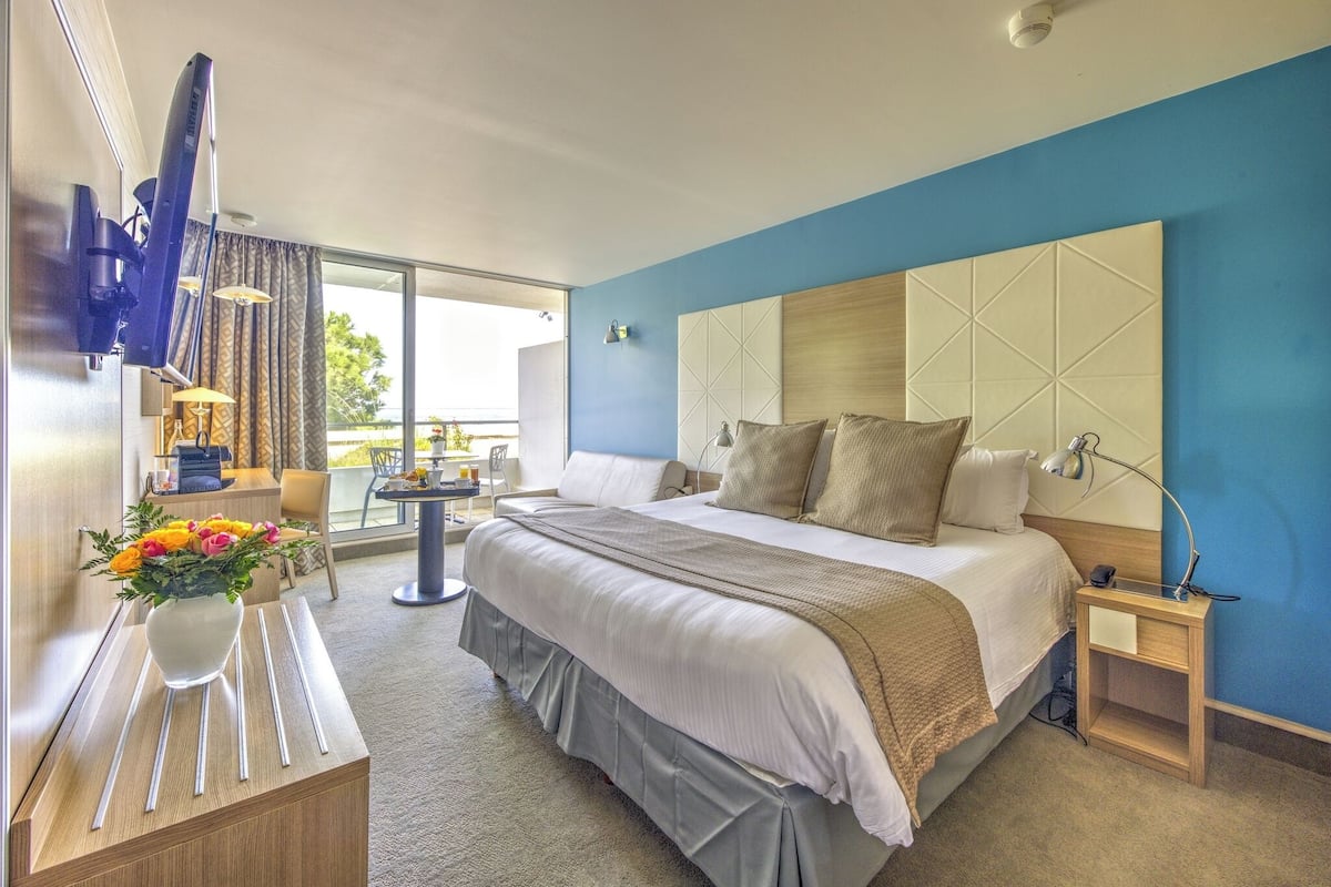 A hotel room with a large bed, a desk with a vase of flowers, a chair, a small table, and a view of the outdoors.