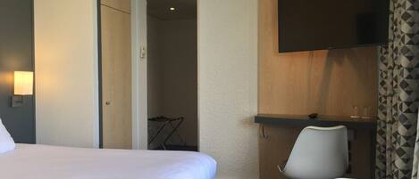 A hotel room with a bed, a desk with a chair, a flat-screen TV mounted on the wall, and a shower area.