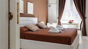 A bedroom with a bed, pillows, a nightstand, a window with curtains, and a view of the outside. - Vatican Garden House (Rome)