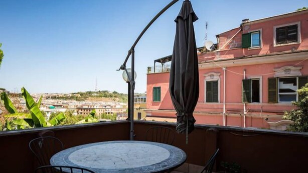 A balcony with a round table and chairs, a hanging umbrella, and a view of a red building with a satellite dish. - Vatican Garden House (Rome)
