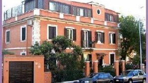 A two-story building with a red facade, a garage, and two cars parked in front. - Vatican Garden House (Rome)