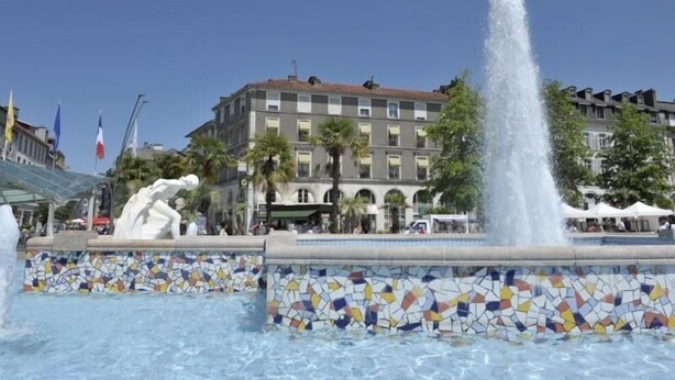 Una fuente con una base de mosaicos, un edificio con múltiples ventanas y balcones, y banderas al fondo.