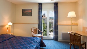 Habitación de hotel con cama, escritorio, silla, ventana con cortinas, radiador y lámpara de pared.