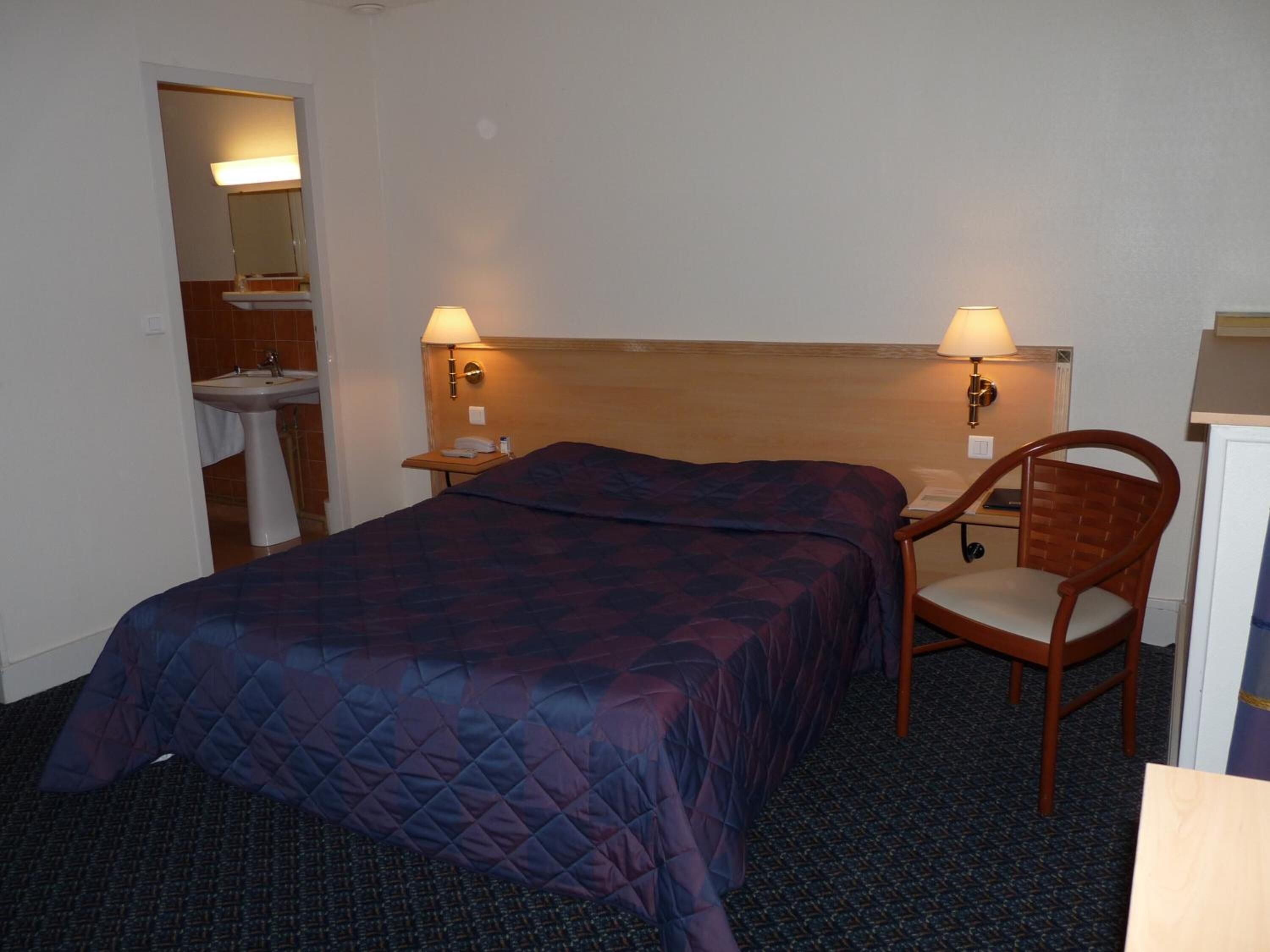 A hotel room with a bed, two bedside lamps, a chair, a small table, a nightstand, and a bathroom visible through an open door.