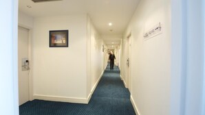 Un pasillo de hotel con alfombra azul, paredes blancas y un cuadro en la pared izquierda.
