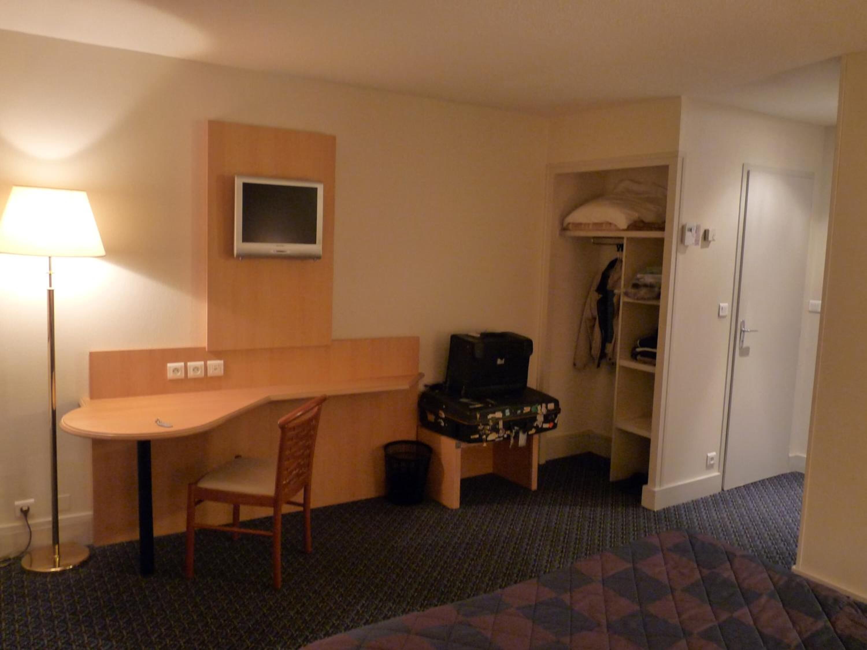 Habitación de hotel con escritorio, silla, televisor, armario y cama con colcha estampada.