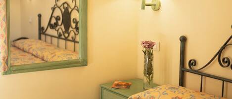 Una cama bien tendida con ropa de cama floral, una mesita de noche con un florero, una lámpara y un espejo en la pared.