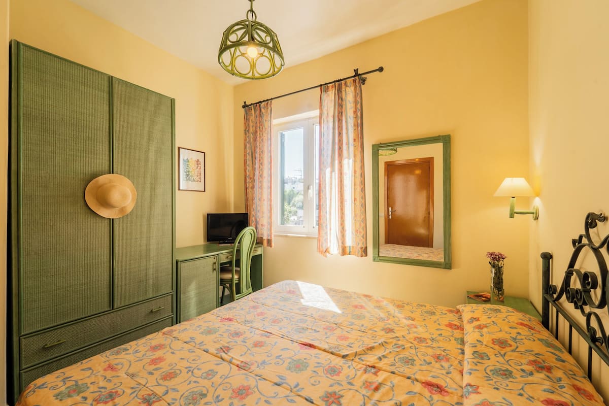 A bedroom with a bed, a desk, a chair, a wardrobe, a television, a window with curtains, and a lamp.