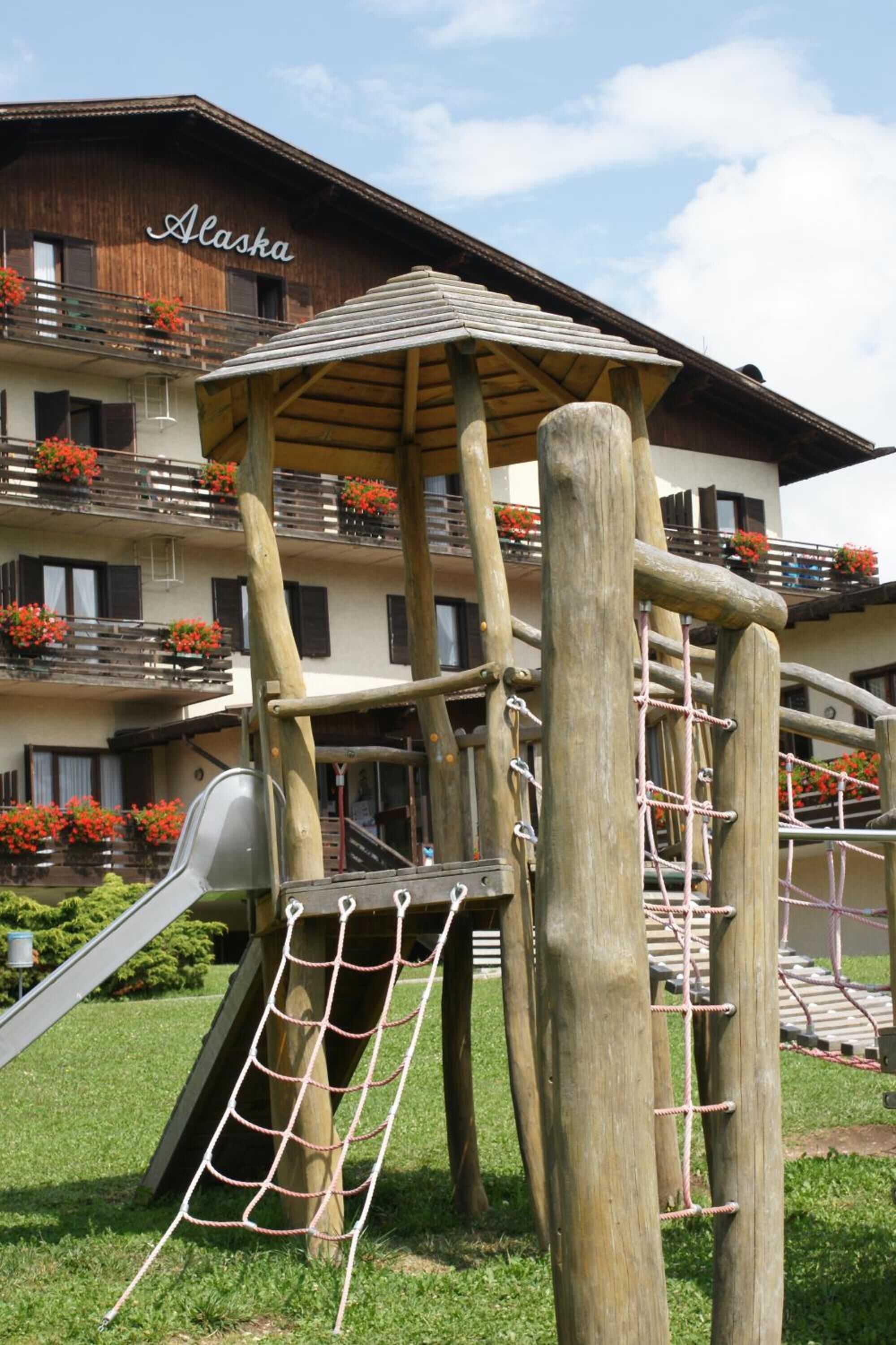 A wooden play structure with a slide and climbing net, set against a building with balconies and flower boxes.