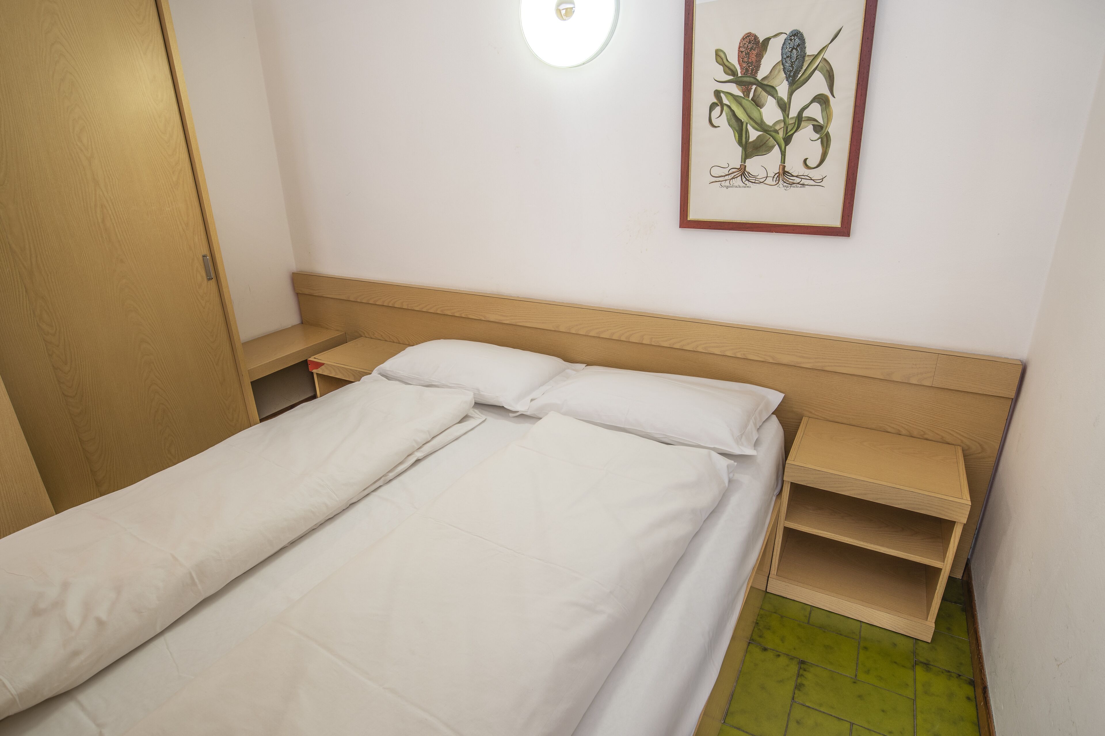 Un lit bien fait, avec du linge de lit blanc, une tête de lit en bois et une petite table de chevet sur laquelle se trouve une lampe.
