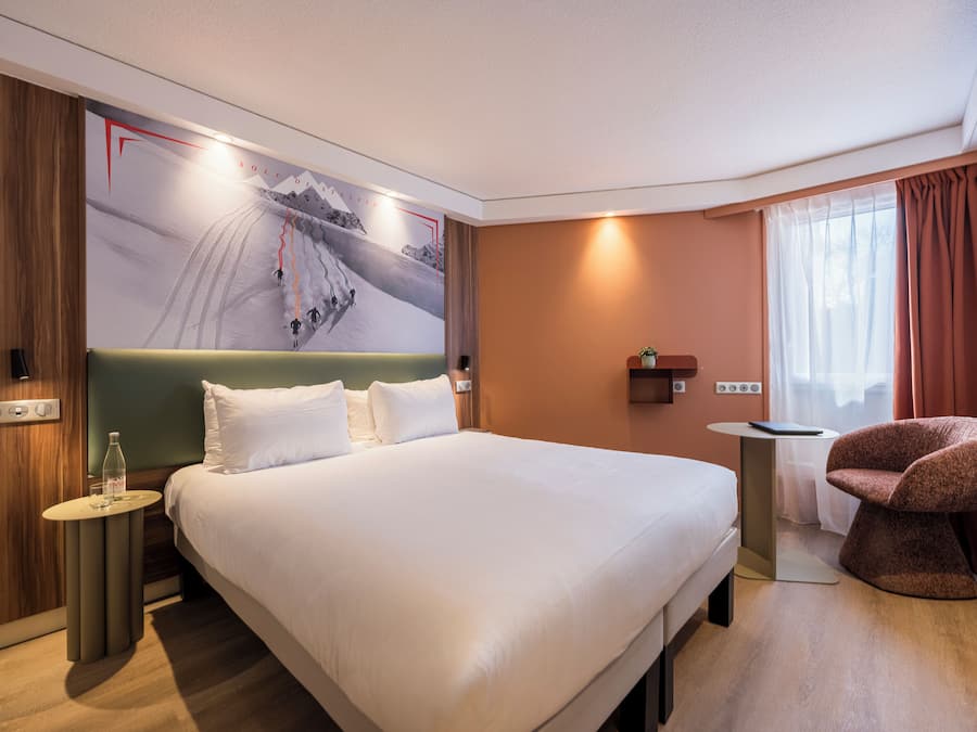 Una habitación de hotel con una cama grande, una mesita auxiliar pequeña, una silla, un estante en la pared y una ventana con cortinas.