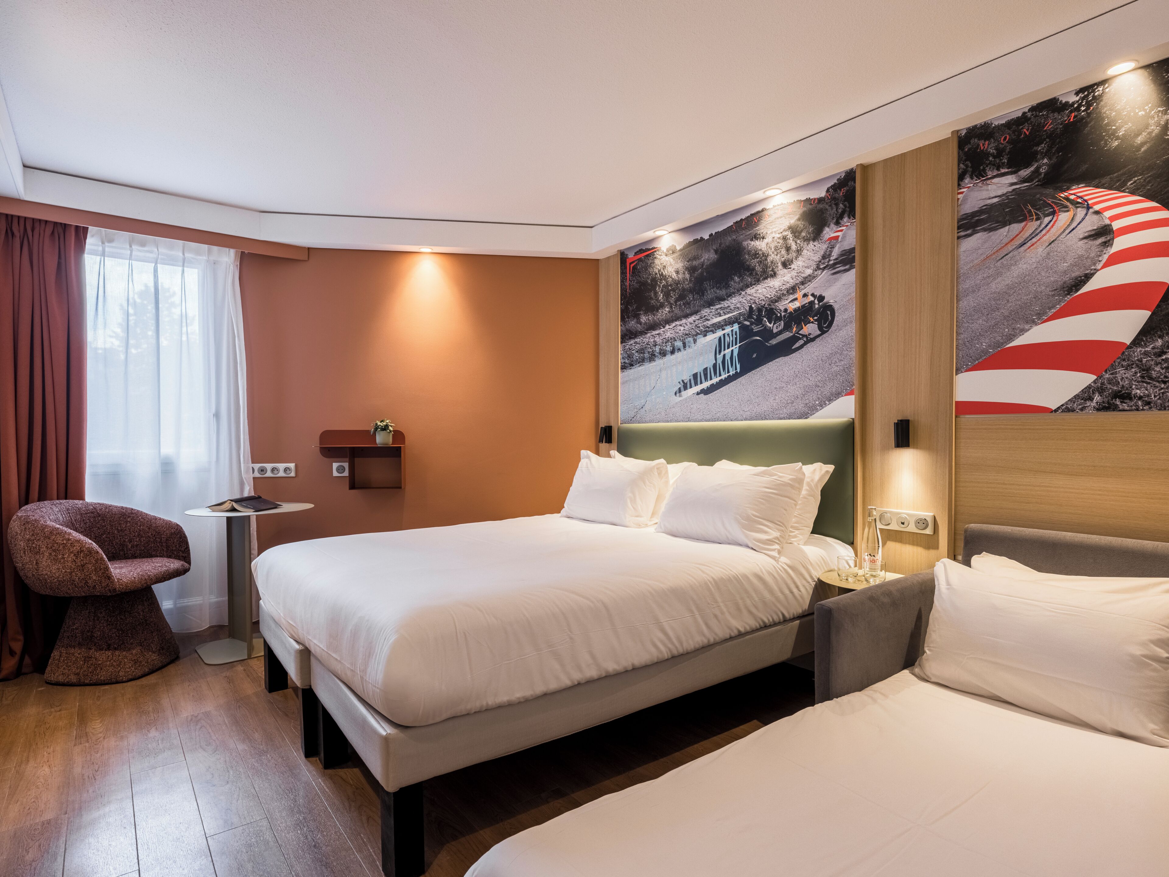 A hotel room with two beds, a small table with a plant, a chair, and a large wall mural.
