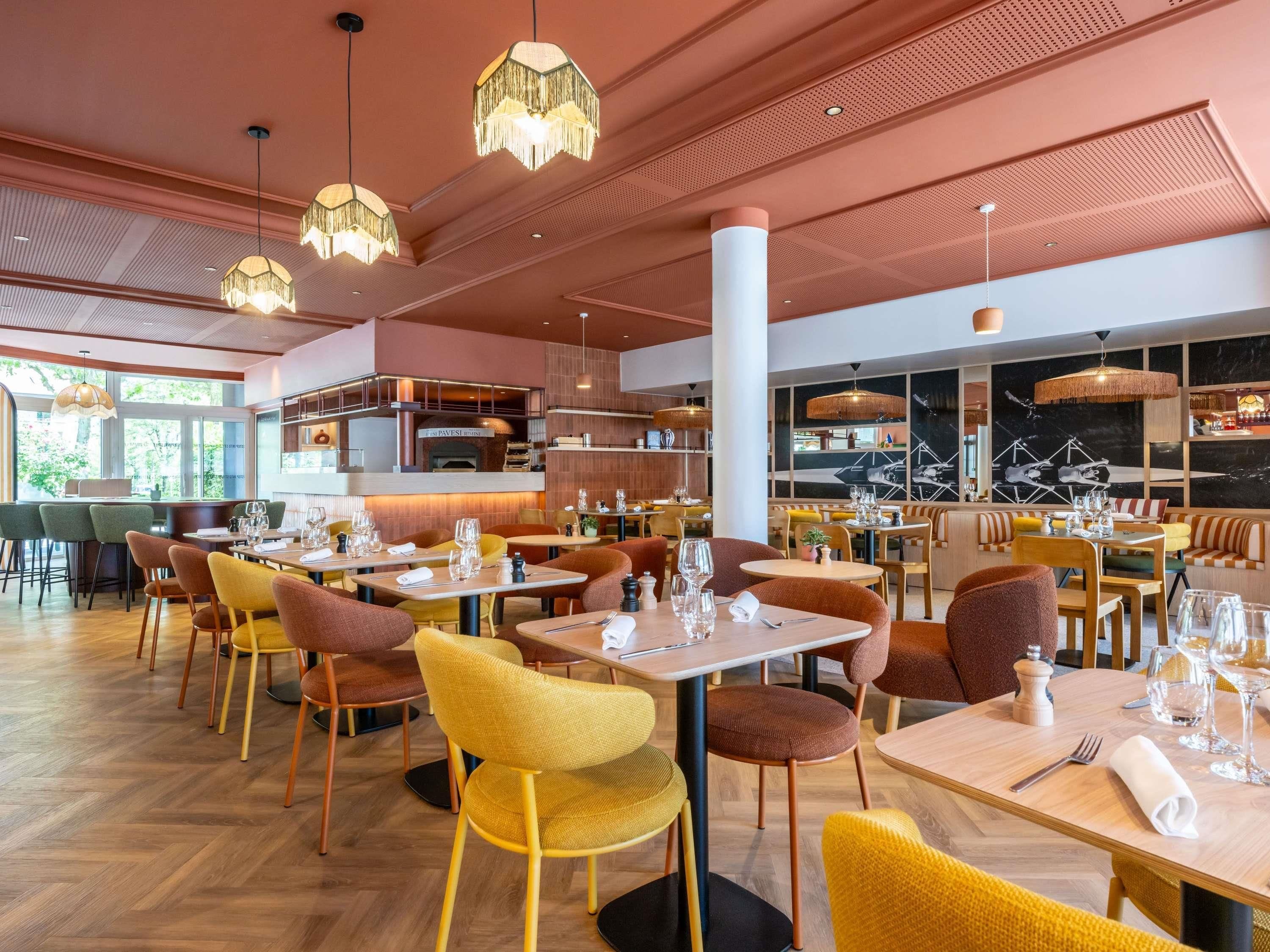 Interior de un restaurante con varias mesas y sillas, una zona de bar y una cocina visible al fondo.