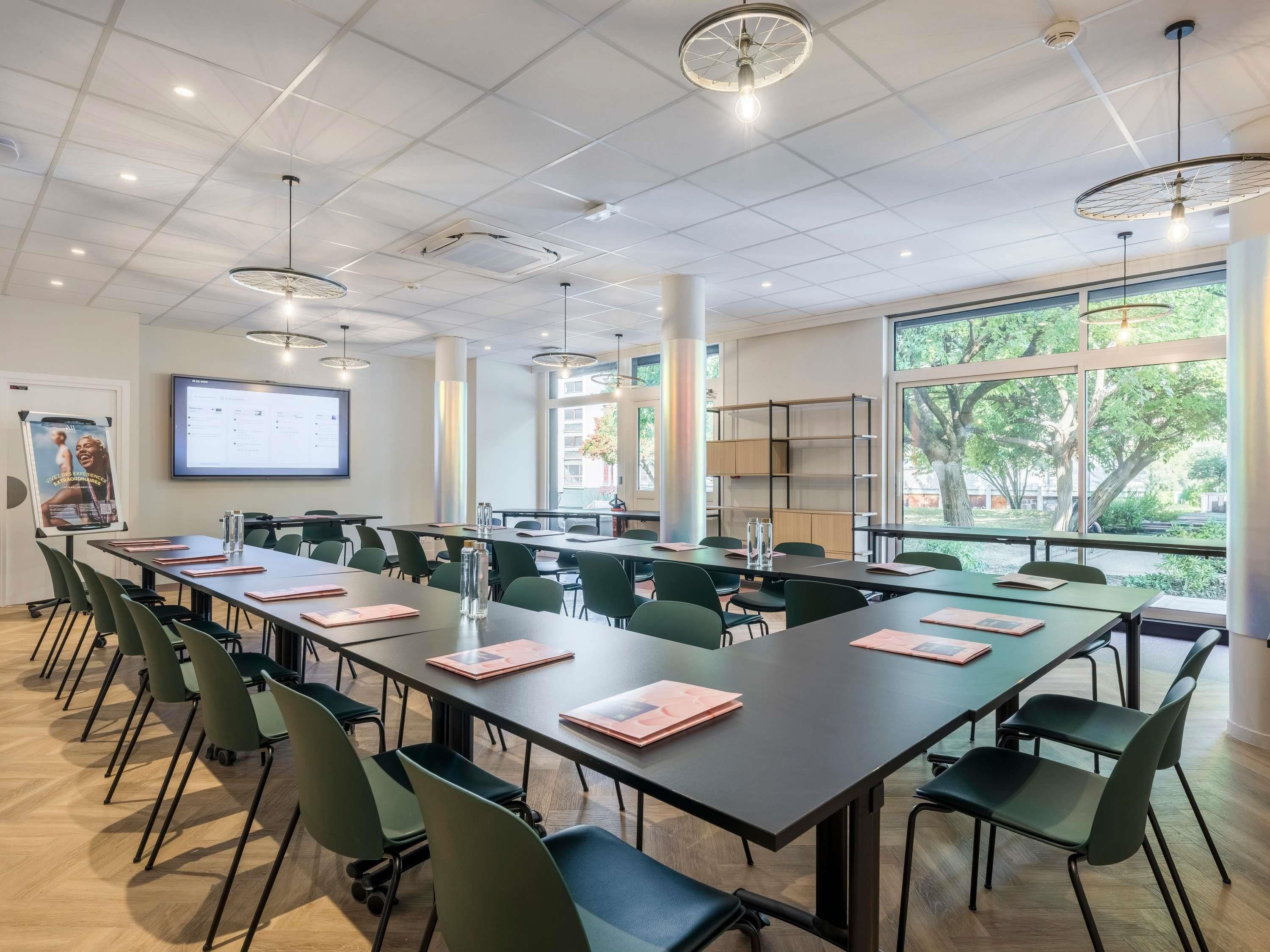 A conference room with tables, chairs, a projector screen, and a large window with a view of trees.
