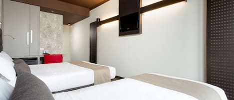 A hotel room with two beds, a red chair, a small kitchenette, and a TV mounted on the wall.